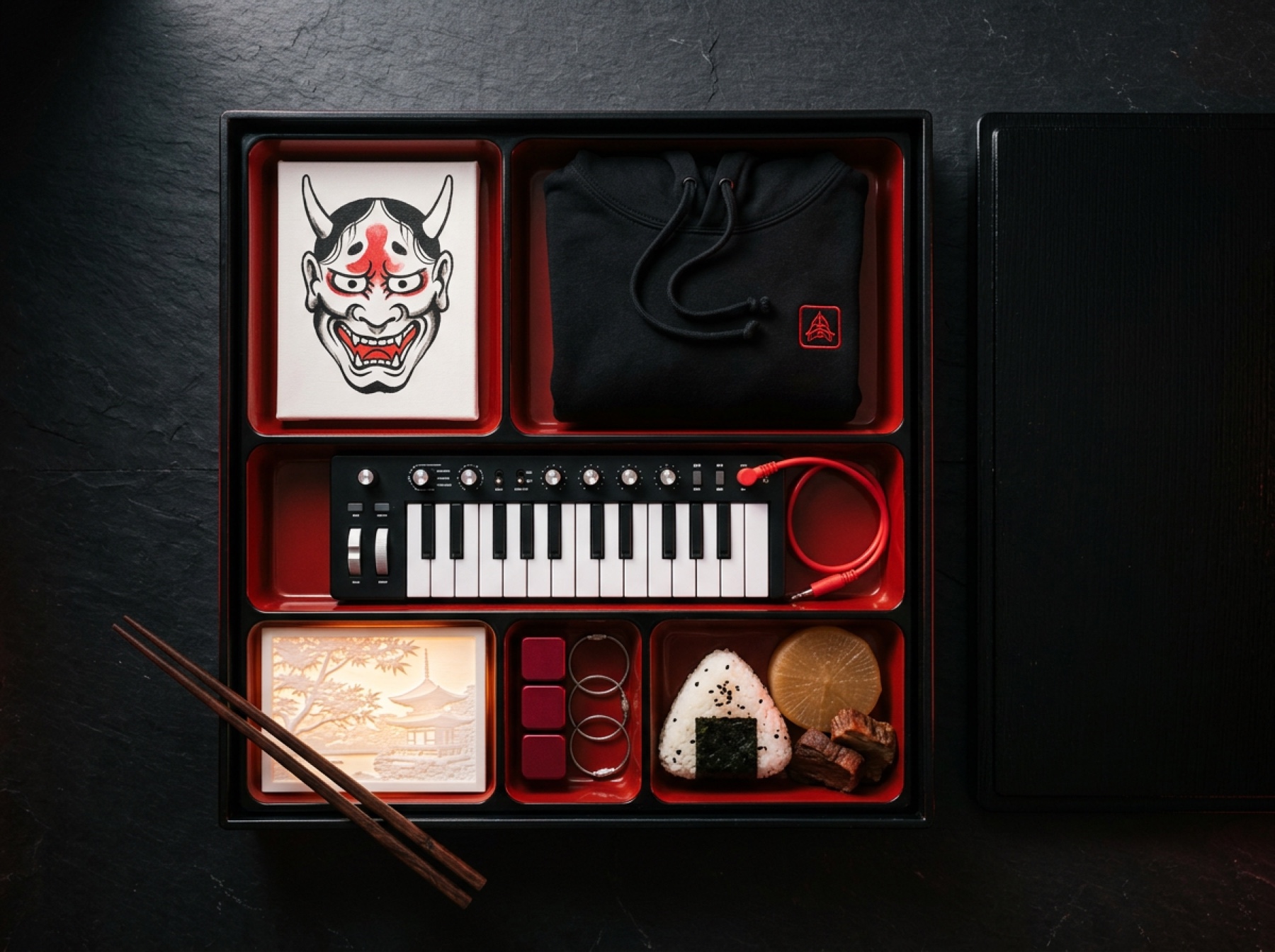 Bento box vista dall'alto: negli scomparti una tela dipinta con un hannya, una felpa nera piegata, un sintetizzatore, una litofania accesa, cubi di alluminio rosso e del cibo vero